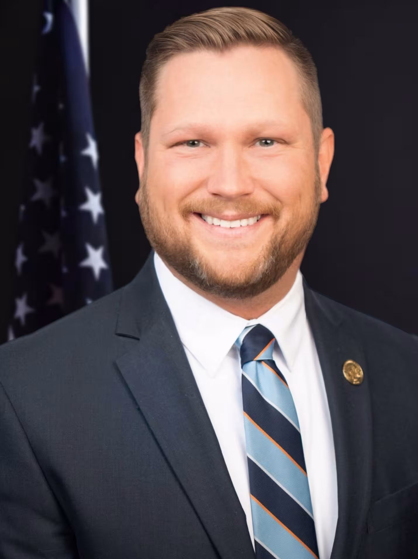 Smiling man in a navy suit and striped tie with a USA flag in the background. He exudes confidence and professionalism.