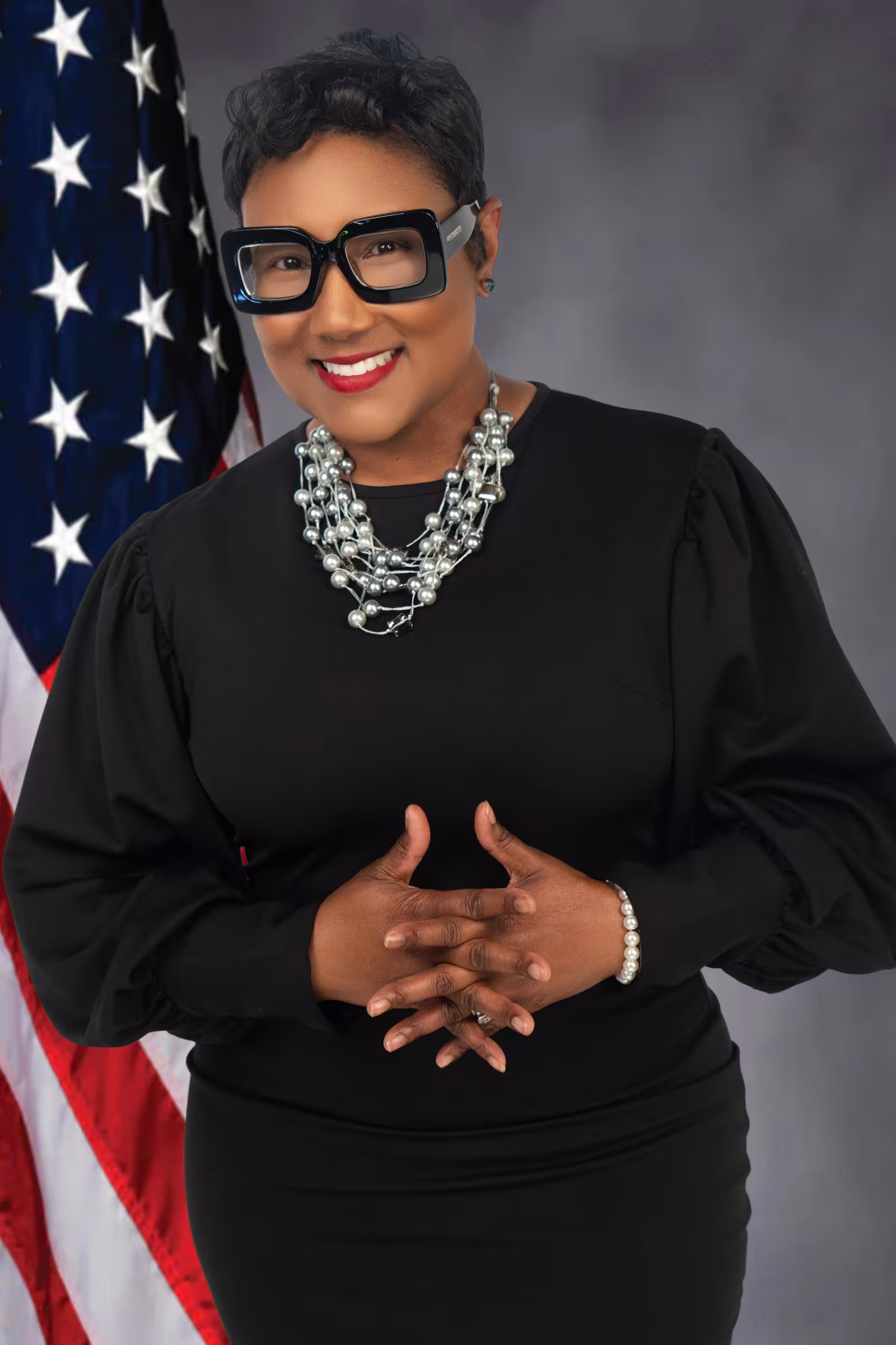 A person smiles confidently, wearing large glasses, a black dress, and a pearl necklace. An American flag is in the background, enhancing the formal tone.