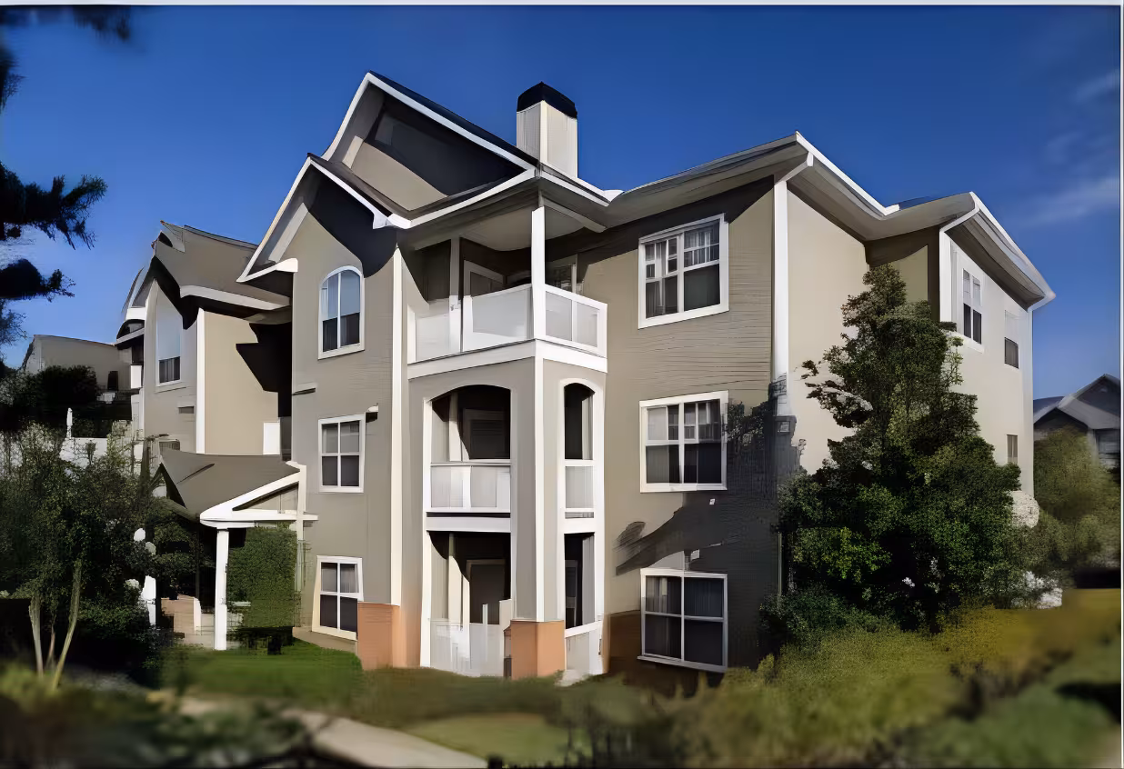 Three-story beige apartment building with white balconies, surrounded by green shrubs and trees, against a clear blue sky. Bright and inviting atmosphere.