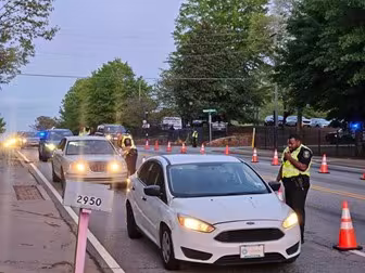Police officers conduct a traffic stop at dusk, wearing reflective vests. Orange cones line the road, and cars wait as an officer checks a driver's details.