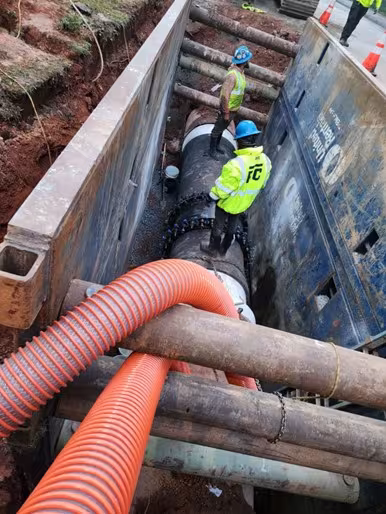 Two construction workers in safety gear stand in a trench alongside a large, installed pipeline. The scene includes protective barriers and orange tubing.