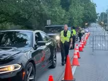 Traffic officers conduct a roadside check on a tree-lined street. Vehicles are queued beside orange cones. The scene is orderly, with a focused atmosphere.