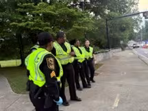 A group of uniformed officers in bright yellow vests stands on a sidewalk near trees, attentively observing an area. The mood is focused and alert.