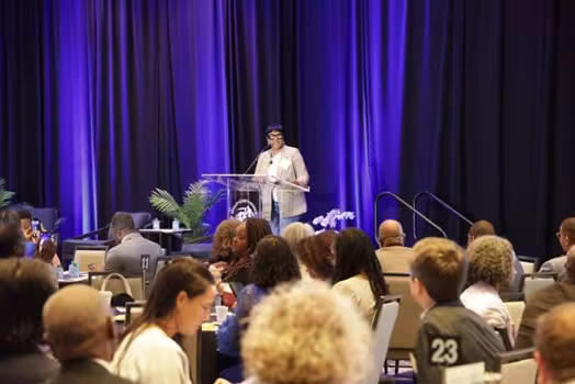 A speaker stands at a clear podium on a stage with blue curtains, addressing a seated audience at a formal event. The atmosphere is professional and attentive.