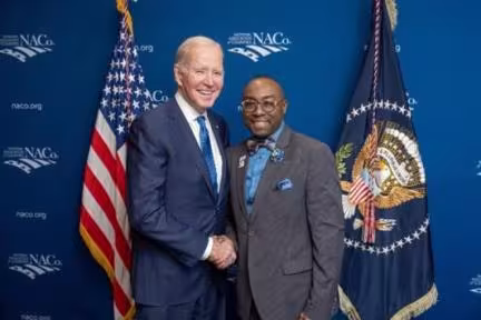 Two men in suits shake hands, smiling against a blue backdrop with flags. The tone is formal and cordial.