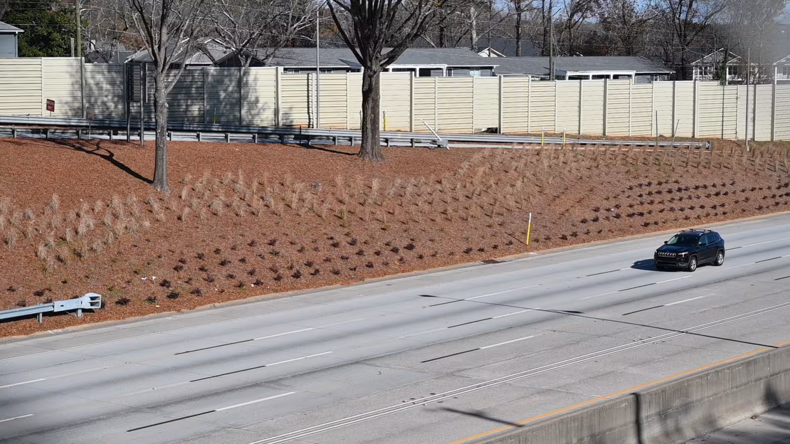 An empty multi-lane road with a single black SUV driving. The backdrop features a sloped, landscaped hill with mulch, young plants, leafless trees, and a beige fence.
