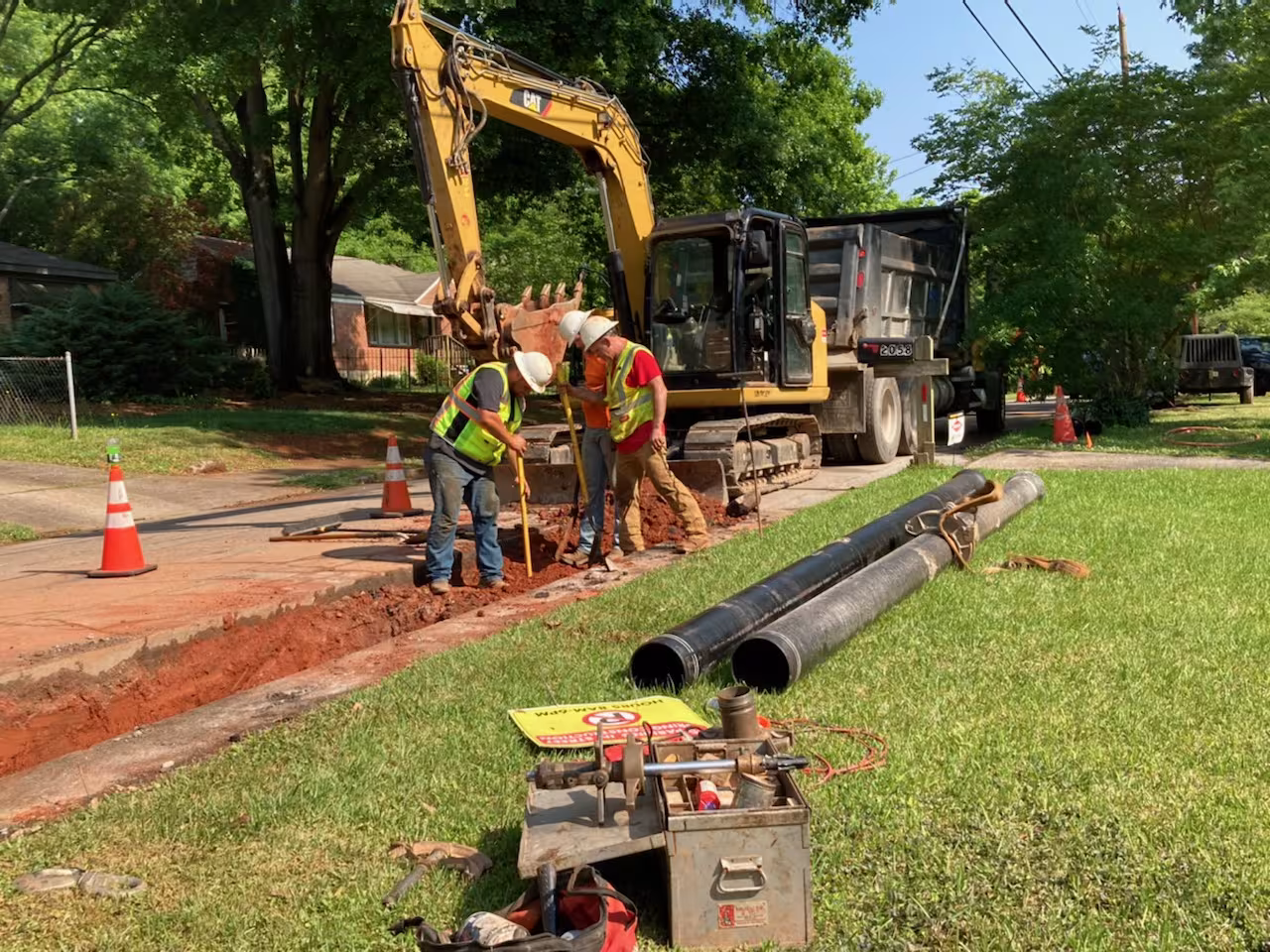 An image of a Caterpillar excavator, a dump truck, and 3 men wearing hard hats digging up a sidewalk with replacement pipes and tools in the grass