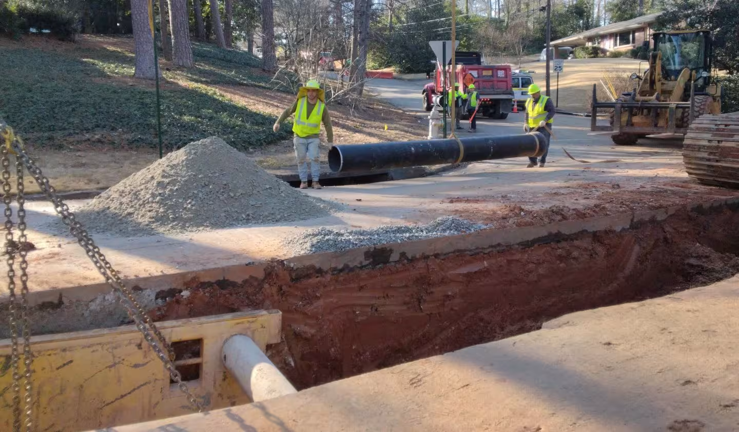 Construction workers in safety vests and helmets work on a road, with a deep trench and large pipes visible. Machinery and vehicles are nearby.