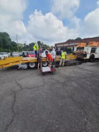Three workers unload metal fencing from a yellow trailer in a parking lot. A red cart is nearby. The scene takes place in front of a building under a cloudy sky.