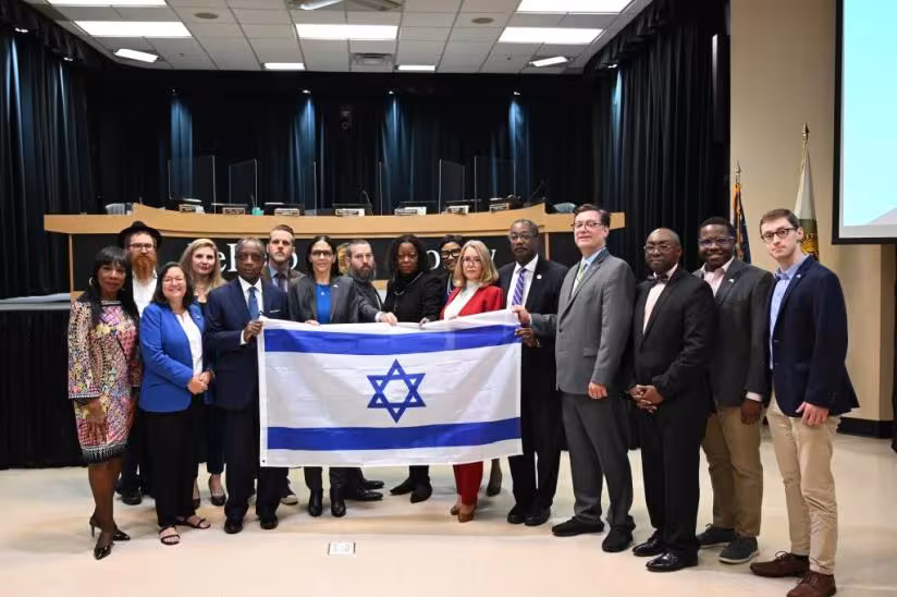 A group of 15 people in business attire stand together in a conference room holding an Israeli flag. They appear formal and unified.