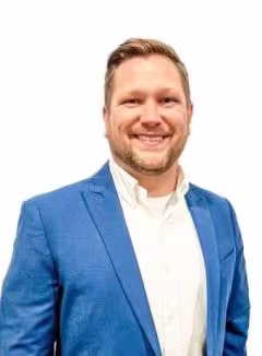 Smiling man wearing a blue blazer and white shirt, standing against a plain white background. He conveys a friendly and professional demeanor.