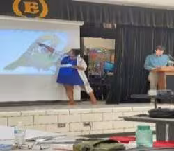 A woman and a man stand on a stage presenting a cartoon bird image on a screen. The setting appears educational, with a lectern and papers.