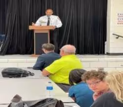 A man speaks at a podium on a stage, addressing an audience seated at tables in front of him. The setting appears to be a community meeting.