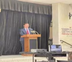 A person in a suit speaks at a podium on a stage with a black curtain backdrop. A laptop on a table faces the audience. The mood is formal and professional.