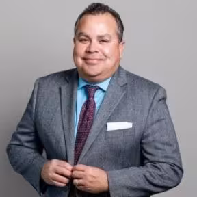 A man in a gray suit, blue shirt, and burgundy tie smiles confidently. He stands against a plain gray background, conveying professionalism and warmth.