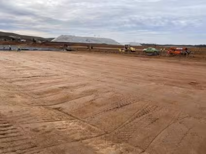 A vast construction site with flattened, reddish earth under a cloudy sky. Vehicles and workers are scattered in the background, conveying a sense of industrious activity.