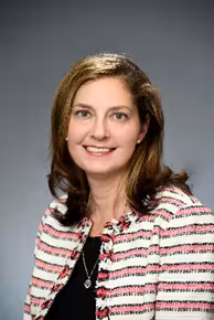 A woman with shoulder-length brown hair and a warm smile wears a striped blazer over a black top. The background is a neutral gray, creating a professional tone.