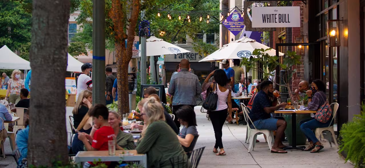 Outdoor dining scene with people enjoying meals at tables under string lights and white umbrellas. Trees and a "White Bull" sign are visible. Casual, lively atmosphere.