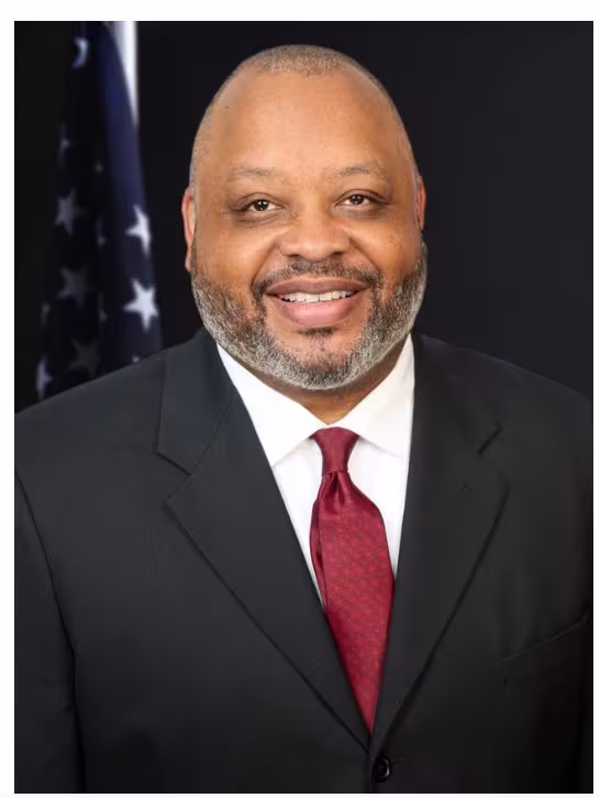 A man with black and grey hair, mustache, and beard smiling wearing a black blazer with a white shirt and wine colored tie. Standing in front of a flag.