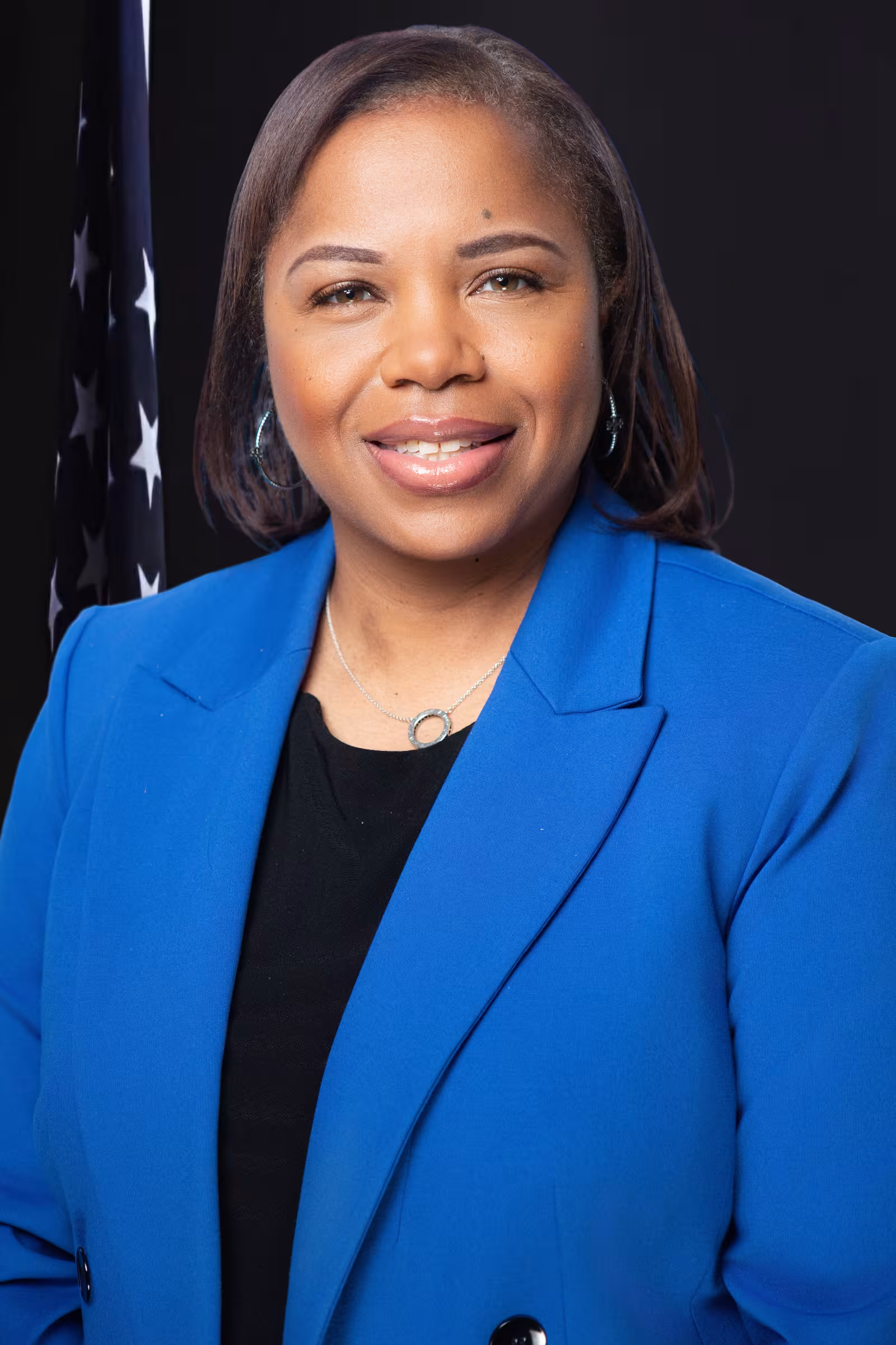 A woman with light brown hair, hooped earrings, and a blue blazer with a black shirt, smiling in front of an American flag with black background.