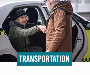 Elderly man wearing a brown coat helping a smiling elderly woman wearing a black hat and green sweater out the back seat of a car.
