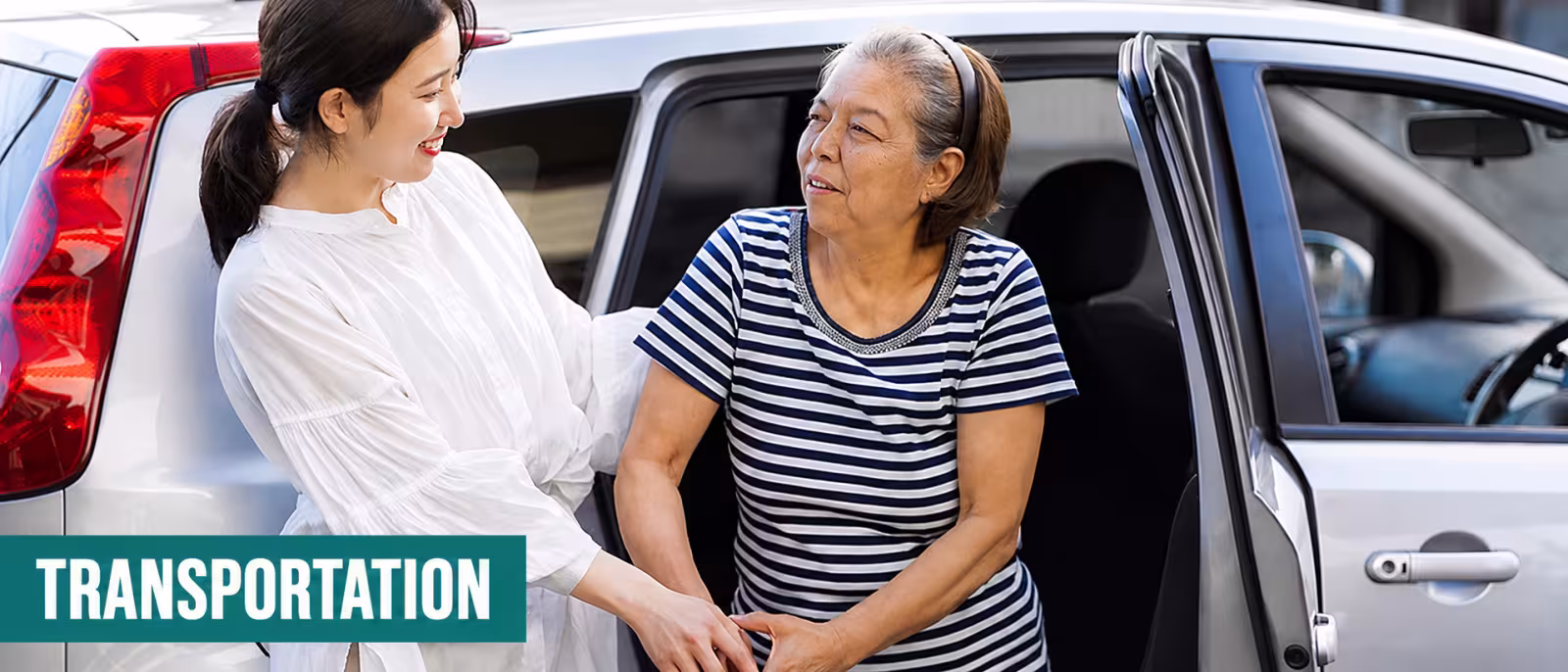 Image of younger woman in white top smiling helping an elderly woman who is smiling wearing a black and white stiped top out the back seat of a silver SUV.