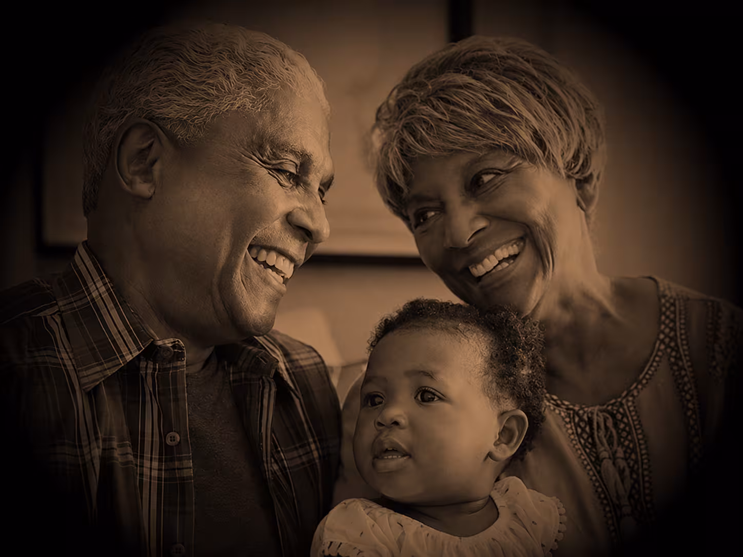 Black & white Image of an elderly man smiling wearing a plaid button up shirt. A baby smiling in the middle wearing a white shirt and an elderly woman on the right smiling with a wall in the background. 