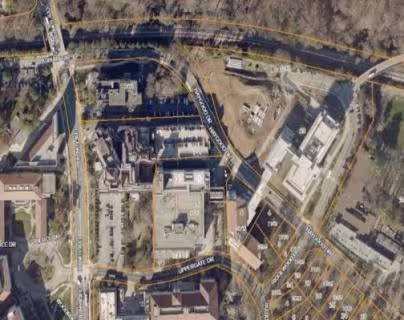 Aerial view of an urban area with a mix of buildings, roads, and greenery. Streets intersect diagonally, surrounded by trees and a small river.