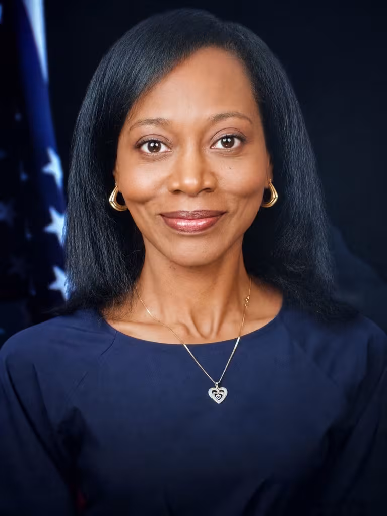 A woman wearing gold earrings with long dark hair smiles warmly, wearing a black blazer and blouse. An American flag is partially visible in the background.