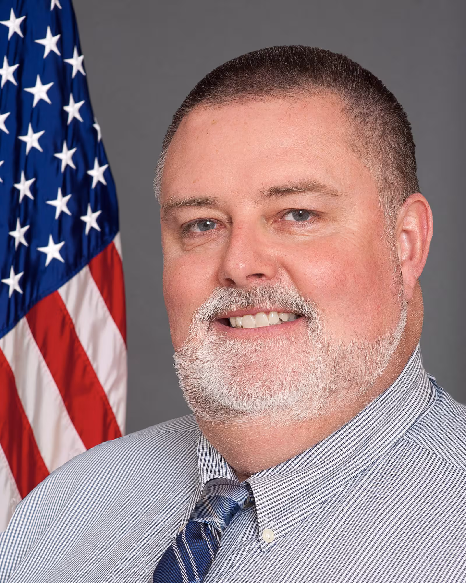 A man in a striped shirt and tie smiles confidently. An American flag is visible in the background, symbolizing patriotism. The tone is formal and professional.