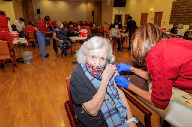 A seated elderly woman receives a vaccination in a community center. Healthcare workers and other recipients are visible in the background. The atmosphere is busy yet organized.