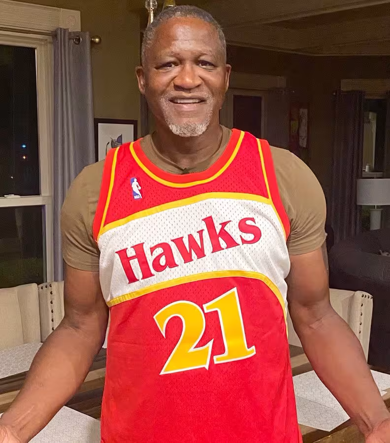 Man smiling, wearing a red and white Hawks basketball jersey with the number 21, stands indoors. The background features a cozy, warmly-lit room.