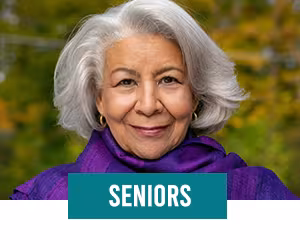 Woman smiling with grey hair wearing a purple top standing in front of greenery landscape