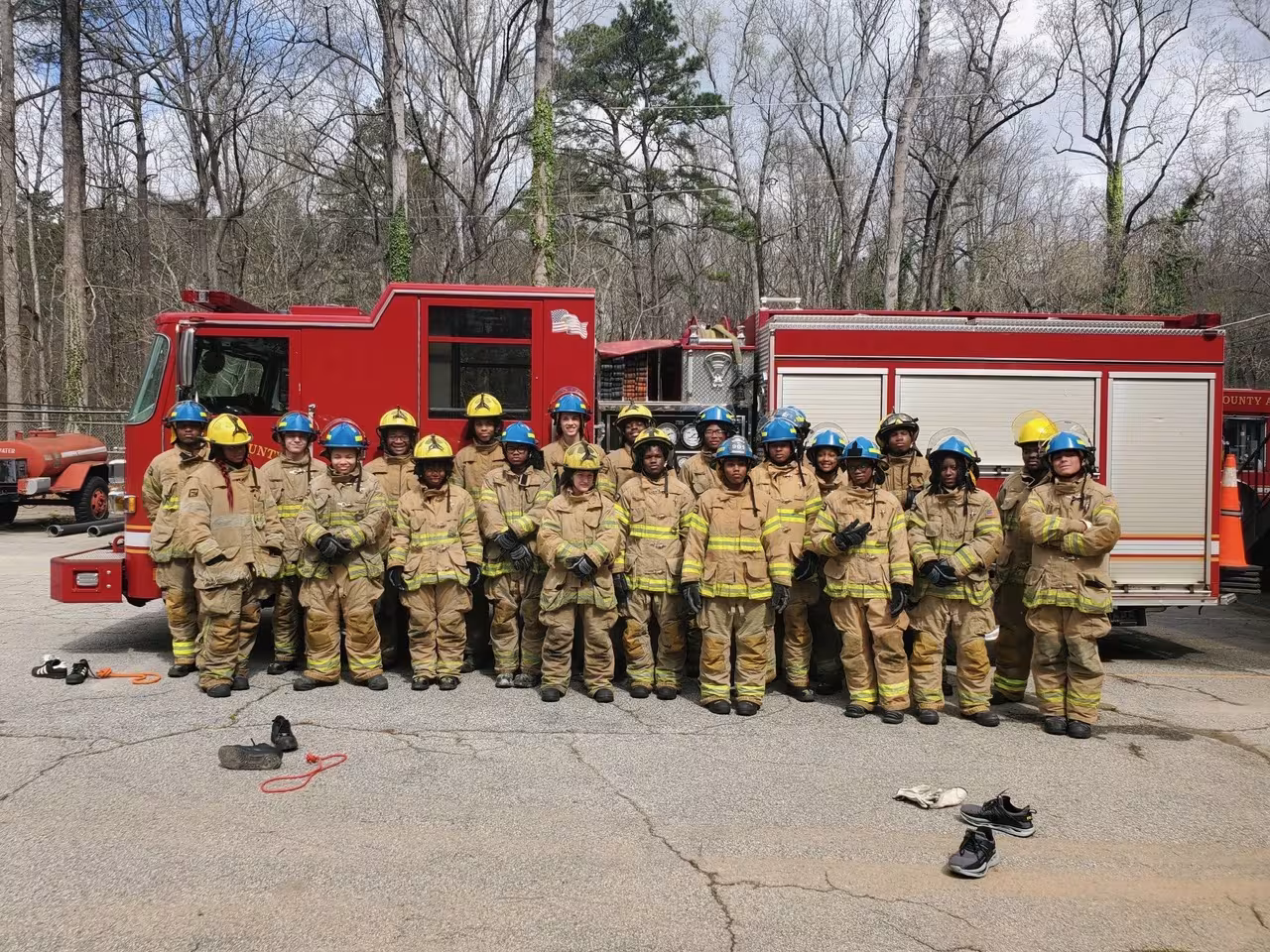 Group of DeKalb County youth firefighters standing in front of a fire truck while gaining hands-on training in full gear with some wearing blue helmets and others wearing yellow helmets. One of them has a black helmet on.
