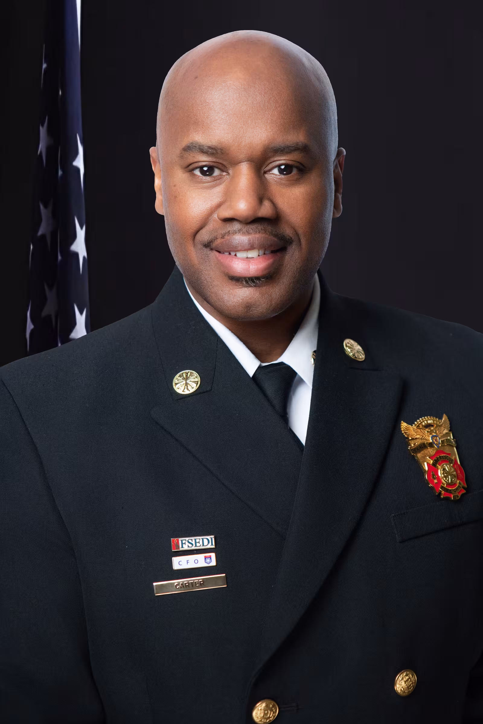 Man in a traditional Fire Rescue dress uniform, a large black suit jacket with a gold badge on it, a white shirt, and a tie, in front of a grey background and an American flag.