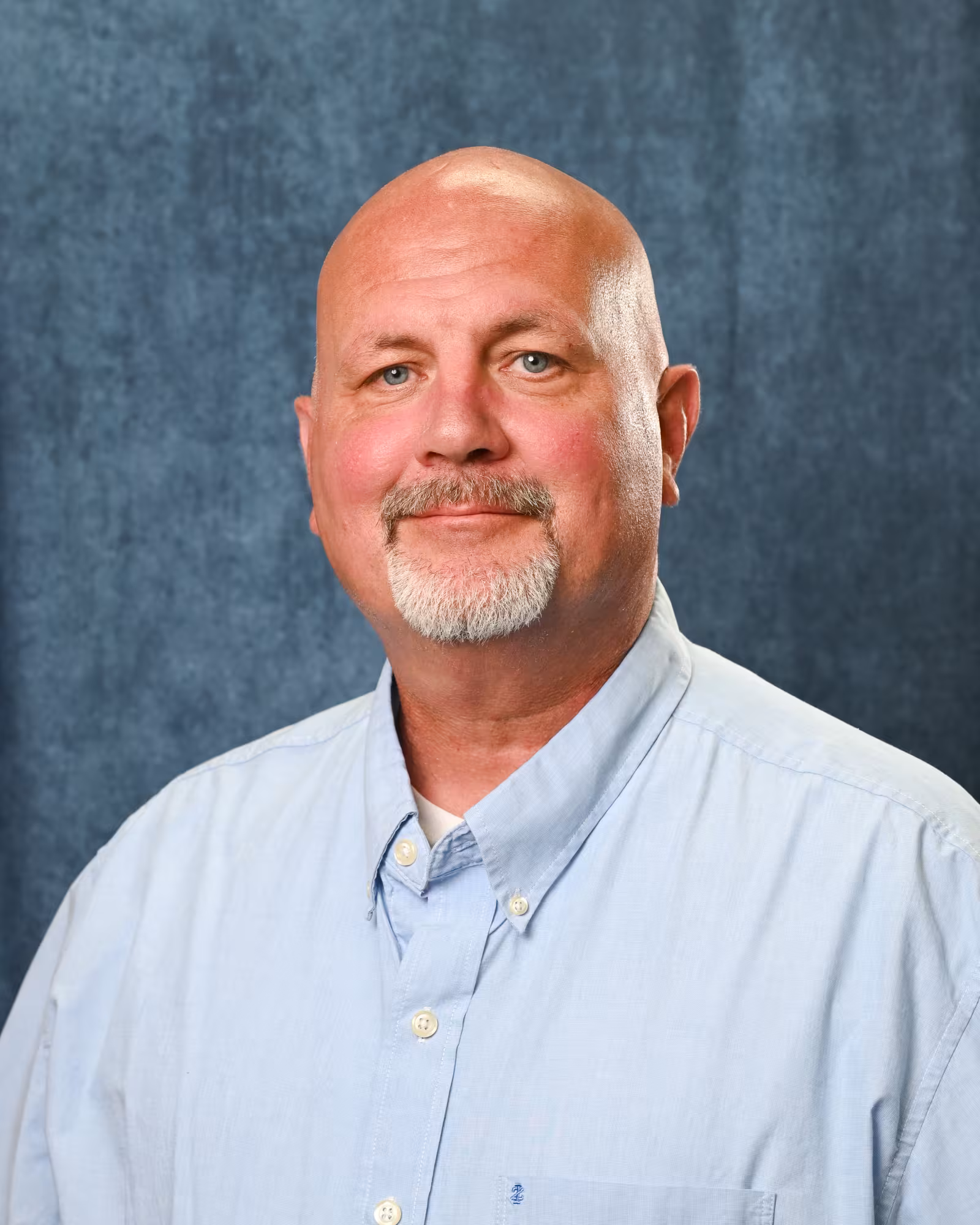 Professional portrait of a bald man with a gray goatee, wearing a light blue collared shirt, in front of a blue textured background.