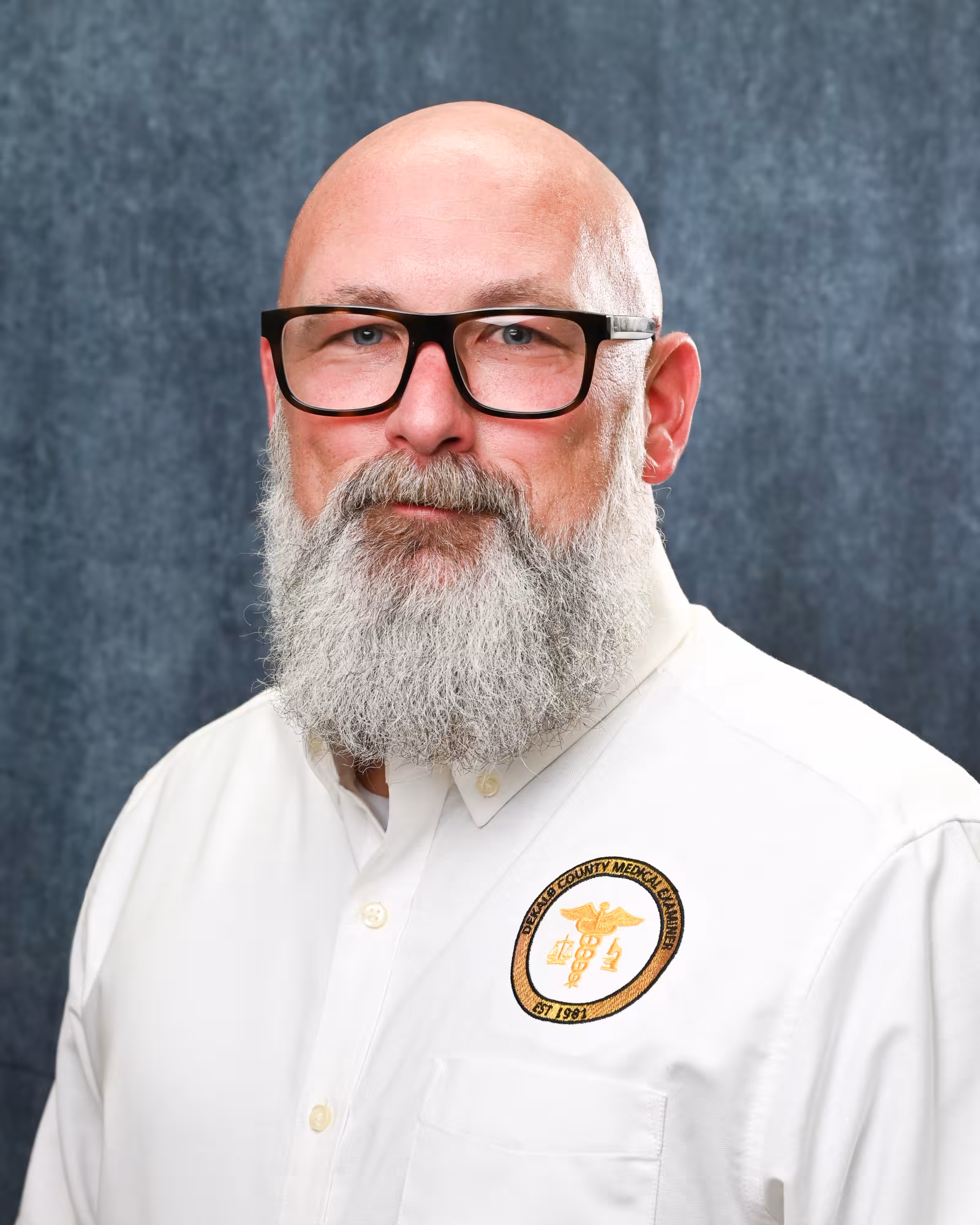Professional portrait of a bald man with a long gray beard and glasses, wearing a white collared shirt with a DeKalb County Medical Examiner patch, in front of a blue textured background.