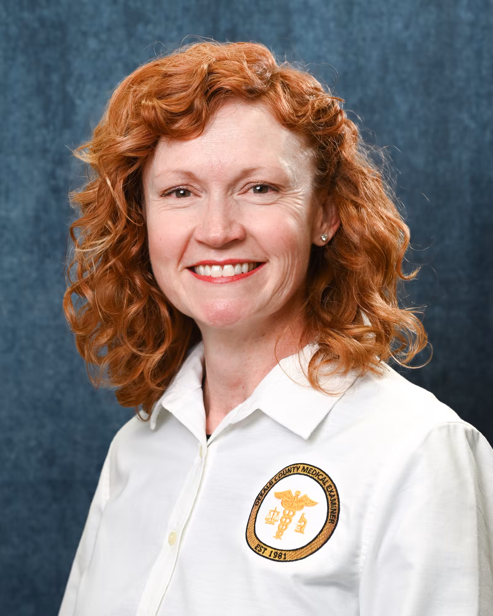 Professional portrait of a woman with curly red hair, wearing a white collared shirt with a DeKalb County Medical Examiner patch, in front of a blue textured background
