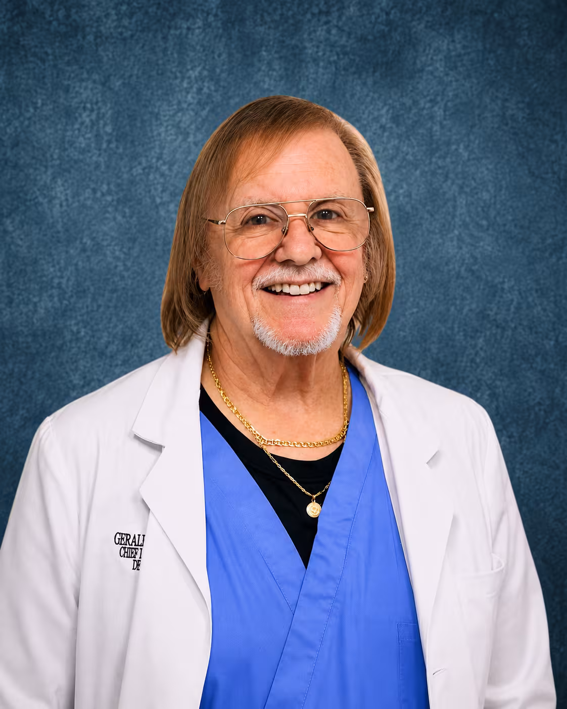 Professional portrait of a man with shoulder-length blonde hair, glasses, and a gray goatee, wearing a white lab coat over blue scrubs, in front of a blue textured background.