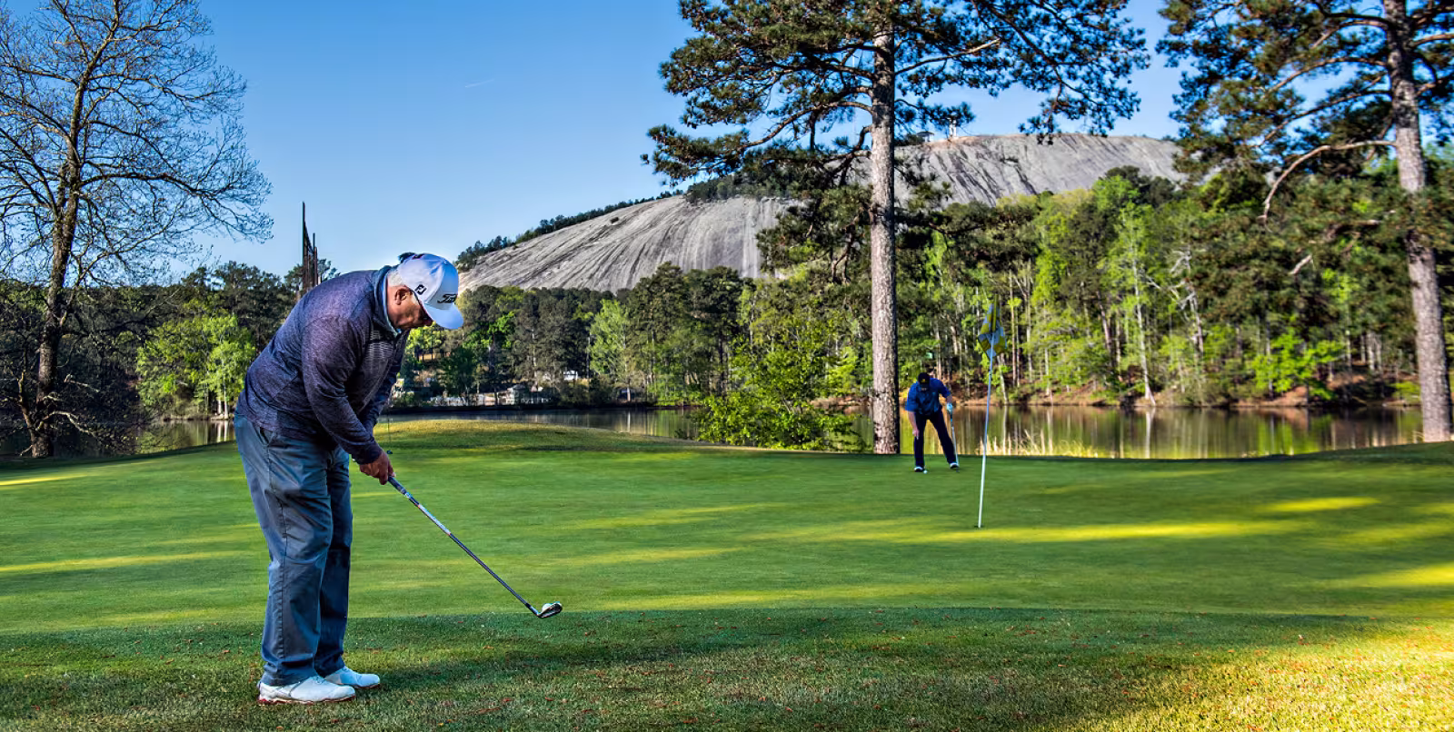 A man in the foreground chipping onto the green while another man stands watching in the background. There is a lake and a mountain in the background and the green is surrounded by trees.
