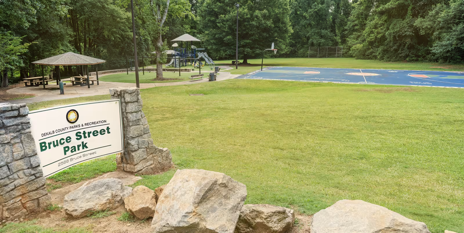 A large green space with a rocky sign in the foreground that reads "Bruce Street Park". There is a picnic pavilion, a playground with a tan covering and a slide, and a blue colored full sized basketball court. The park is surrounded by green trees.