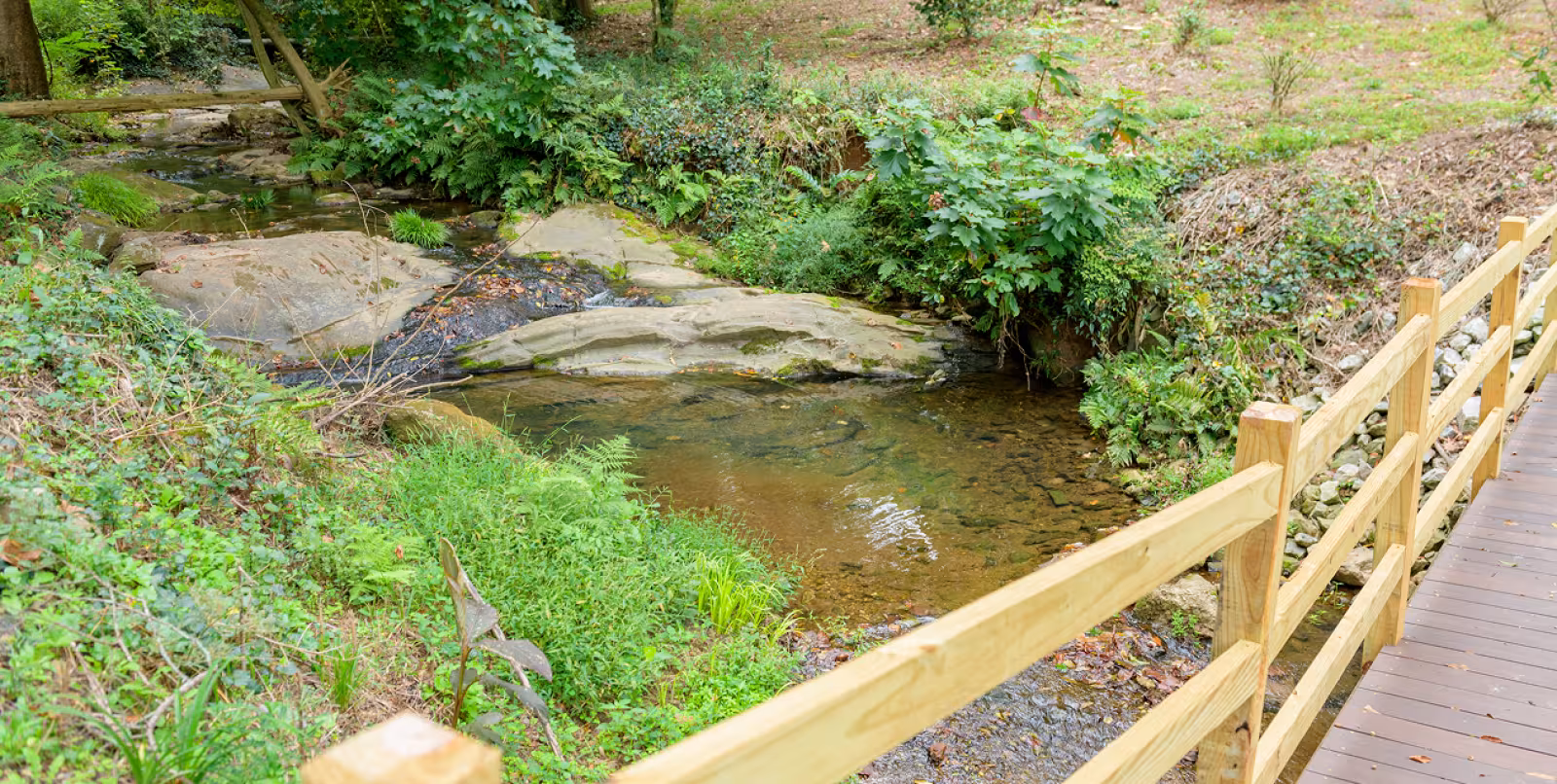 A bridge going over a small green with water flowing down several large rocks surrounded by greenery.