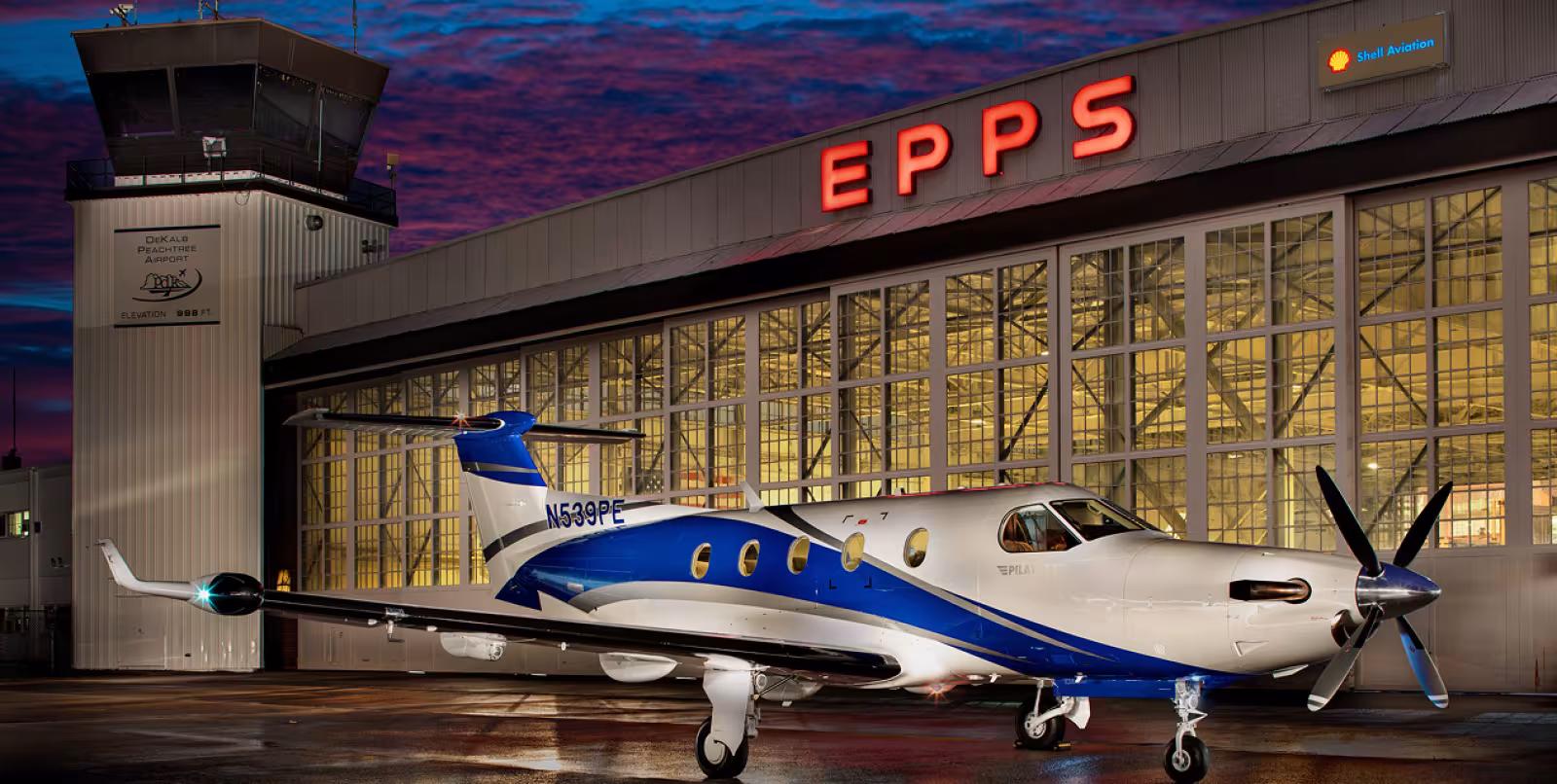 Small white plane with blue accents parked in front of a large hanger with the word "EPPS" in large red letters above the hanger at night.