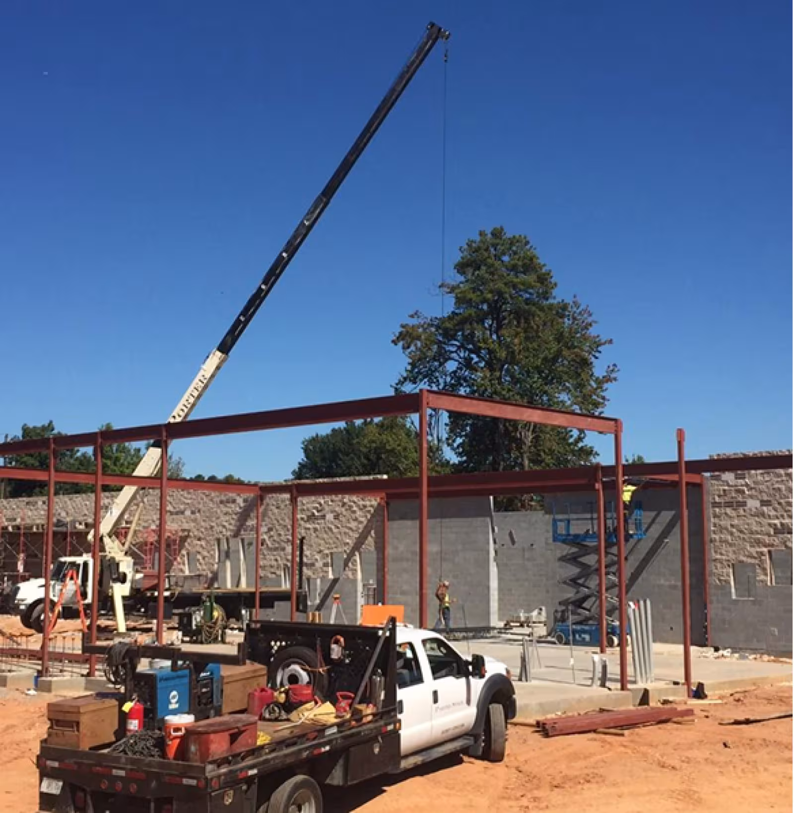Construction site with a metal structure, a concrete floor, and surrounded by dirt. There is a truck with equipment on the back and a crane in the blue skied background.