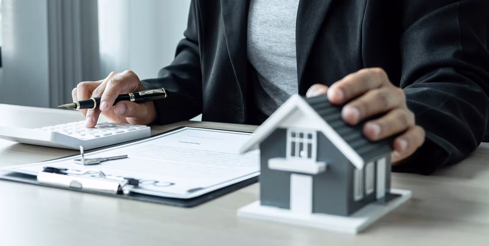 Person sitting at a desk punching in numbers on a calculator with a form on a clipboard and a hand on a grey and white model of a basic home.