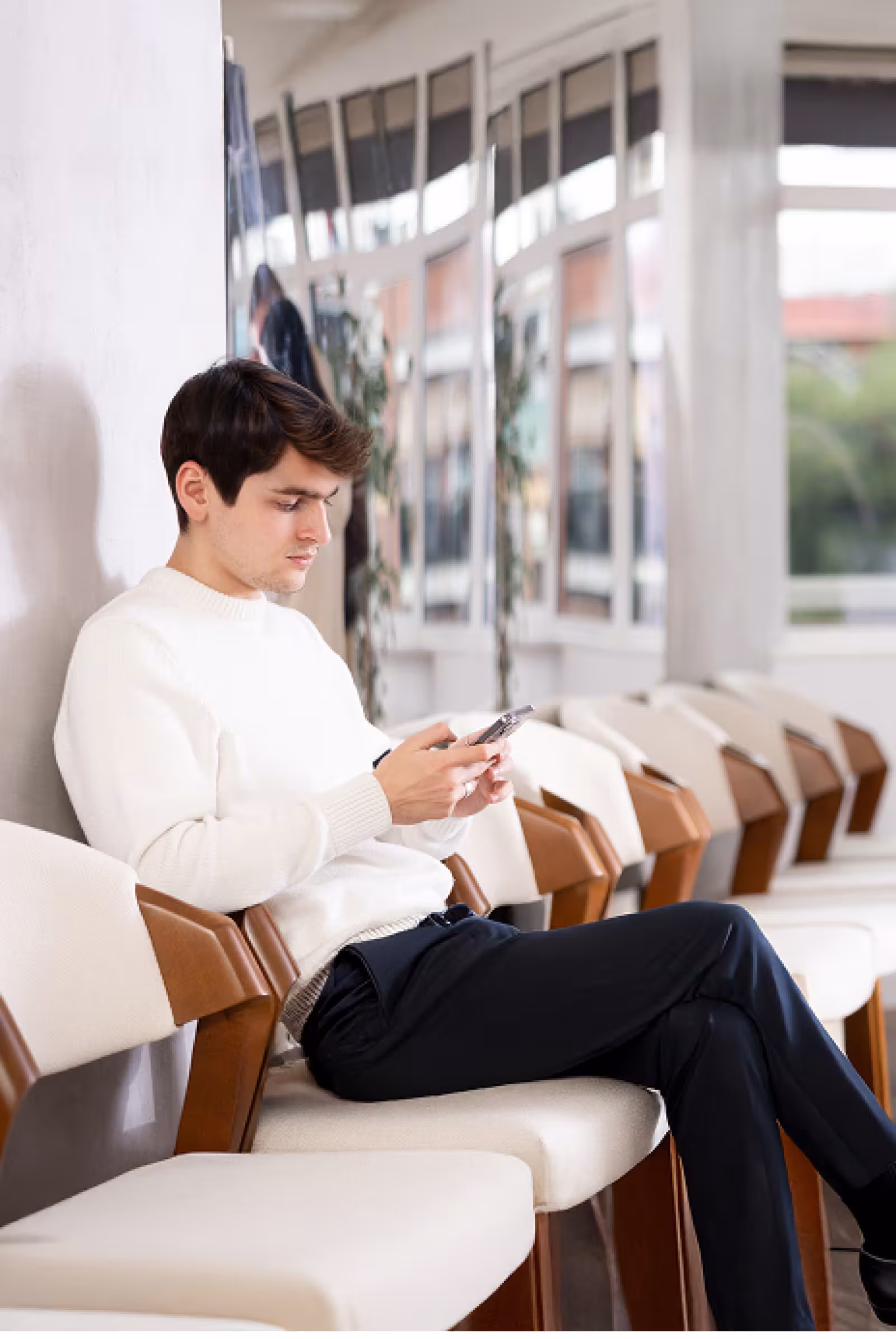 Man siting by himself in a white and wooden chair in a white walled waiting area on his phone. He is in a white shirt and black pants.