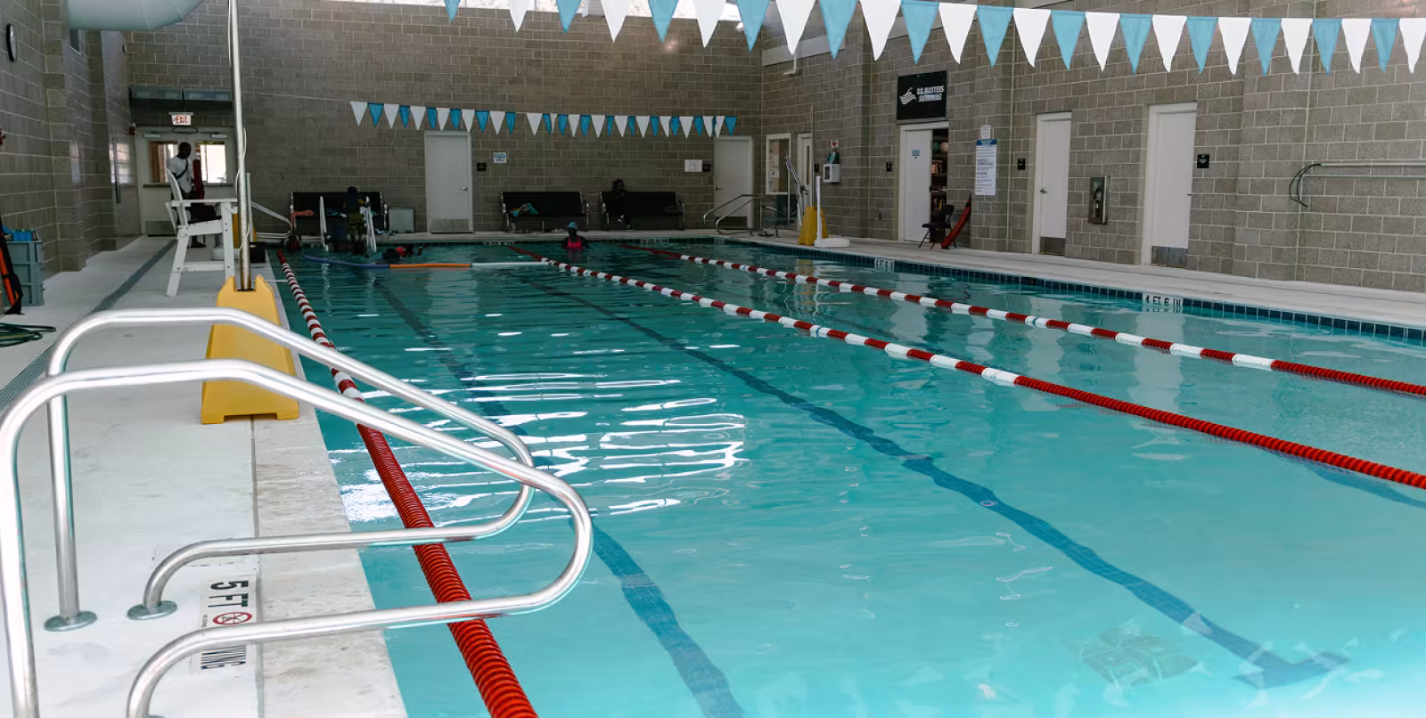 Indoor swimming pool with clear blue water, red lane dividers, and blue triangular flags above. A few swimmers and lifeguards are present. Bright, clean ambiance.