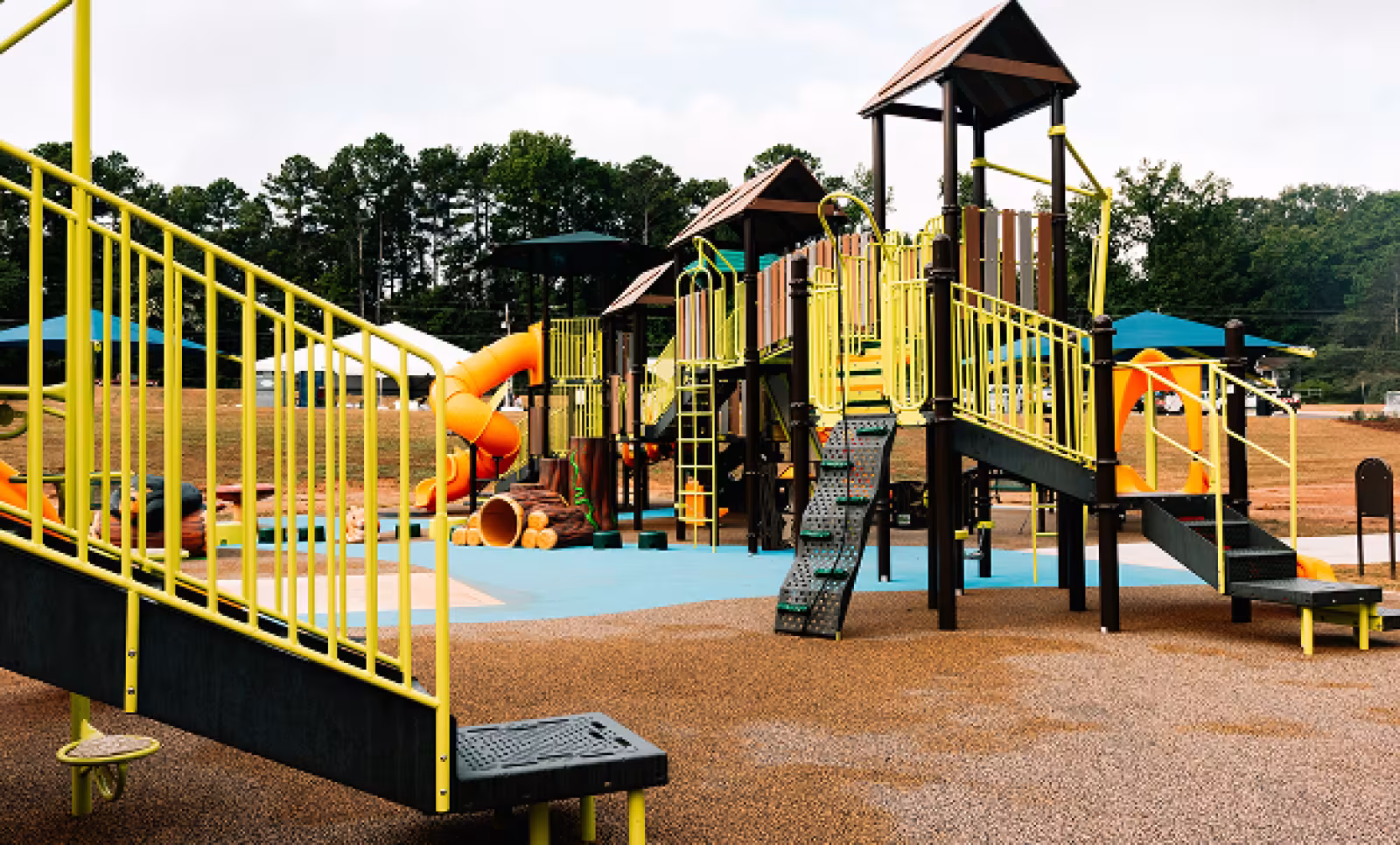 A colorful playground with yellow railings, slides, and climbing structures is set in an open area with trees in the background, conveying a lively and inviting atmosphere.