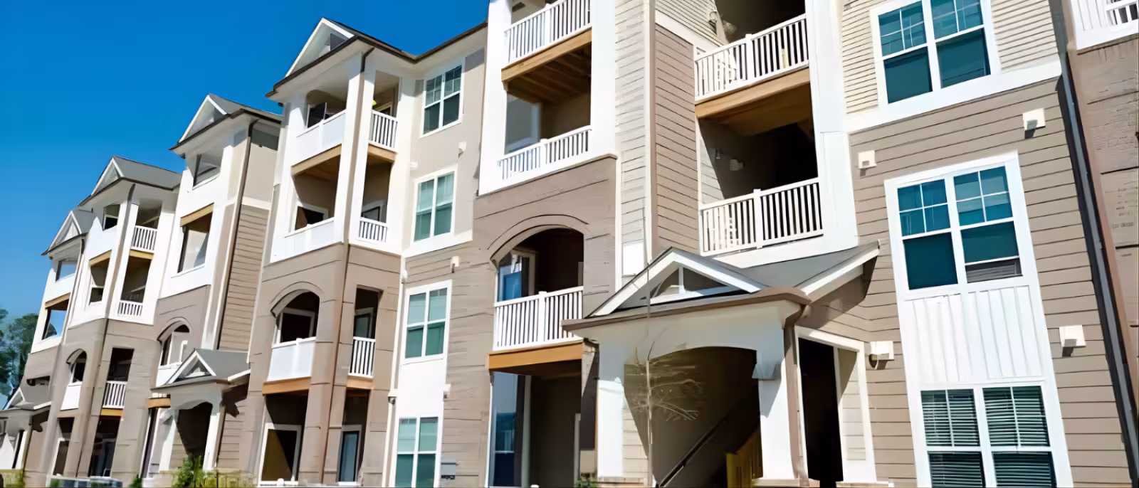 Modern apartment building exterior with multiple floors, balconies, and large windows. The facade features beige and white tones, under a clear blue sky.