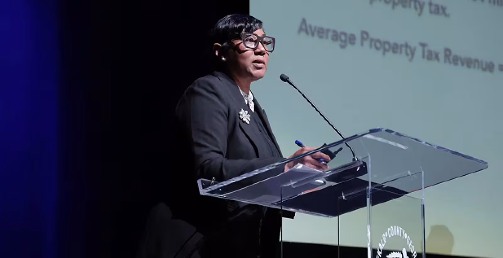 A person in a suit speaks at a podium against a dark background, displaying a presentation on property tax revenue. The setting is formal and professional.